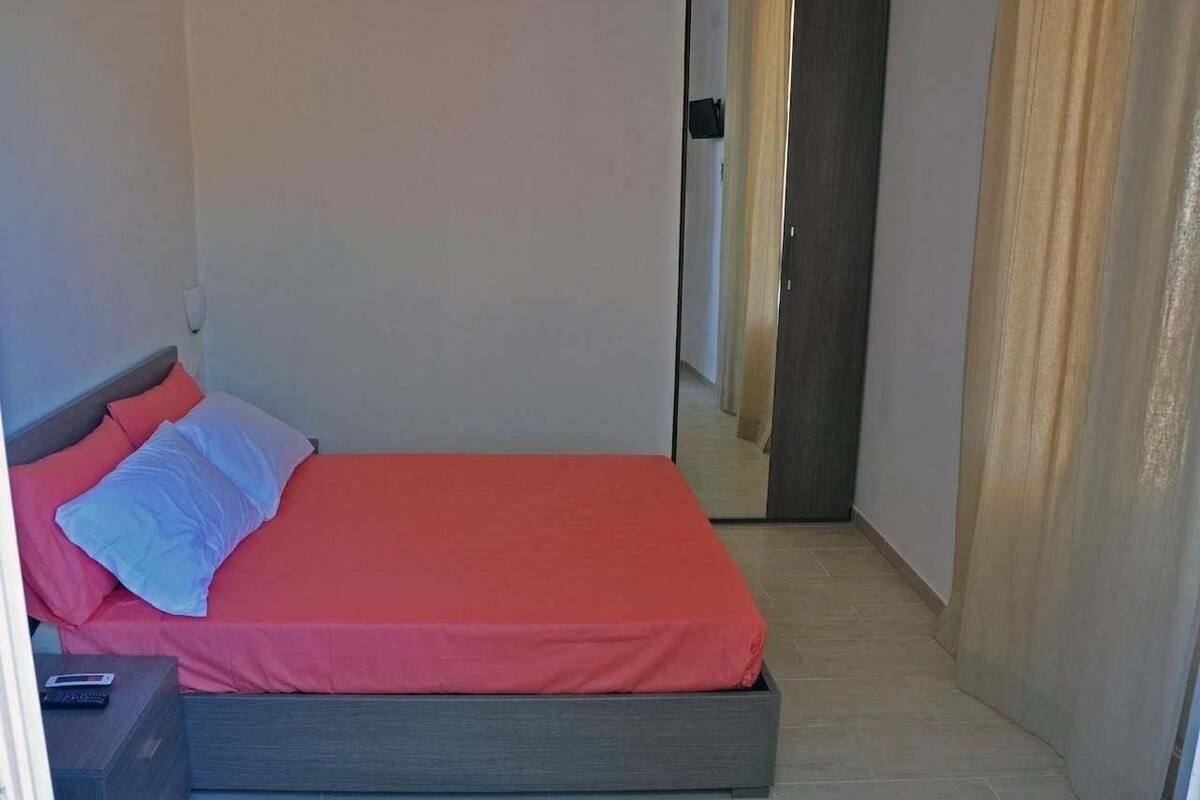 A small, narrow room with a single bed, a nightstand, and a door leading to another room.