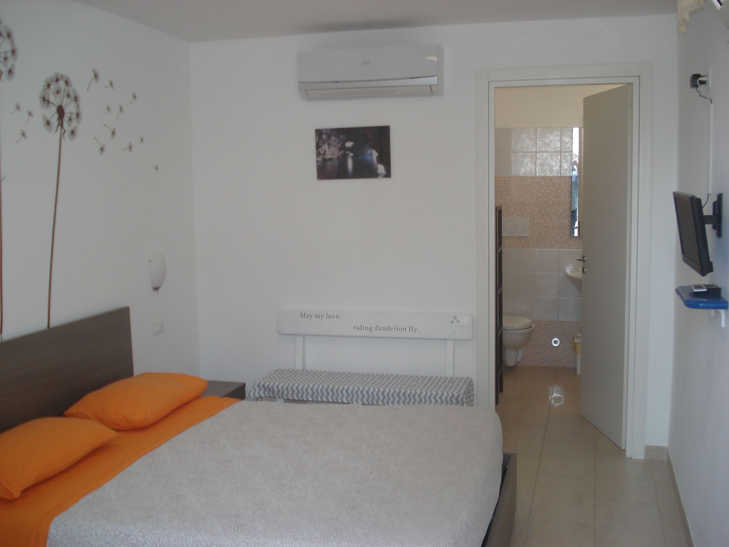 Un dormitorio con cama, aire acondicionado, un cuadro y un baño visible a través de una puerta abierta.