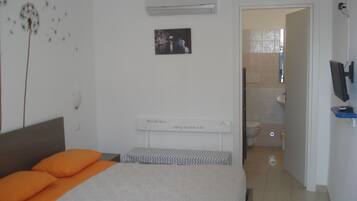 Un dormitorio con cama, aire acondicionado, un cuadro y un baño visible a través de una puerta abierta.