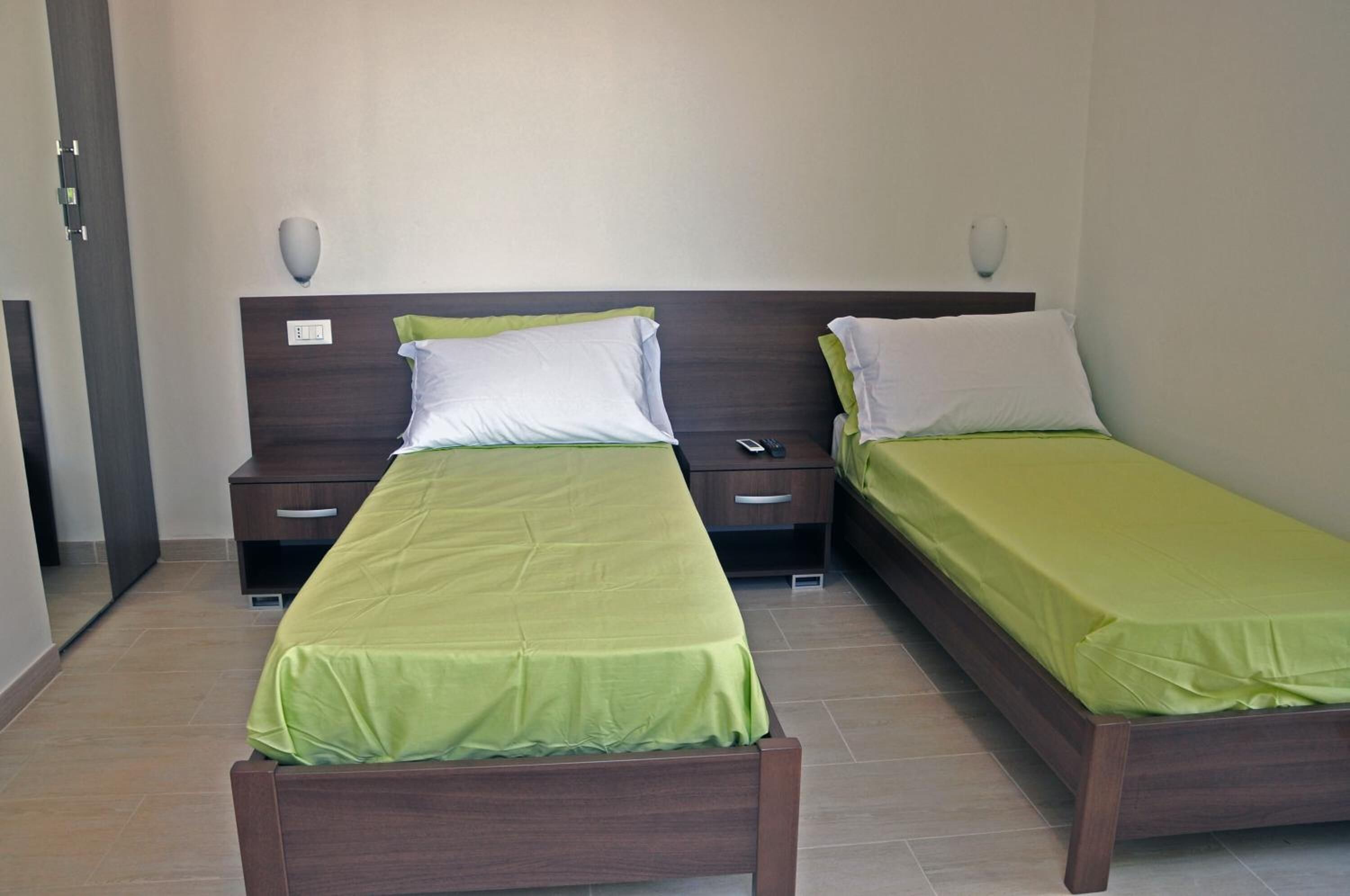 Habitación de hotel con dos camas individuales, cada una con cabecero de madera y ropa de cama verde. Hay mesitas de noche con lámparas y un enchufe en cada cama.