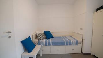 Ein kleiner, weißer Raum mit einem Bett, einer Sitzbank und einem Kleiderschrank.