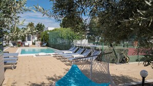Zona de piscina con sillones de descanso, una hamaca y una cancha de tenis al fondo.