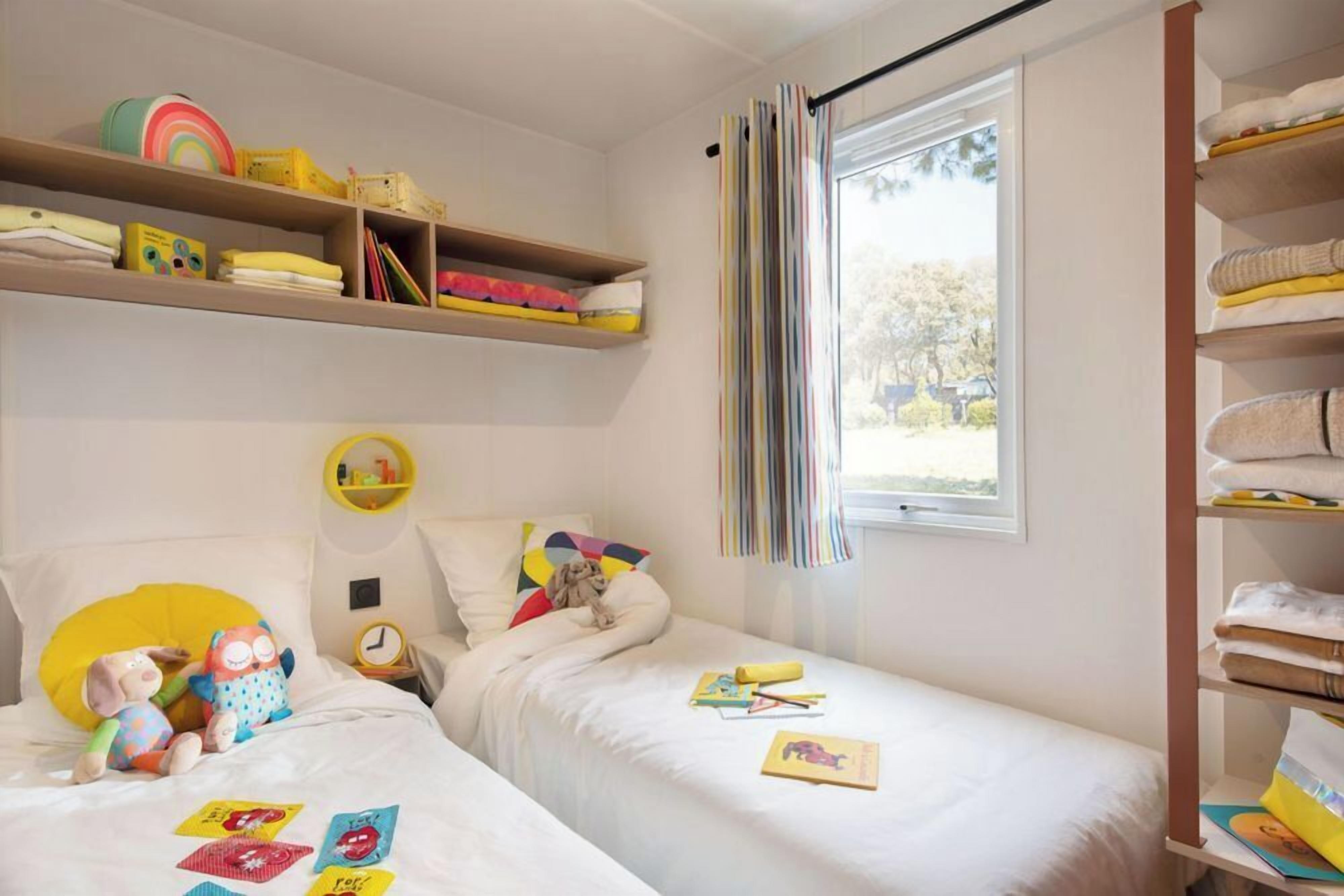 Quarto de criança com duas camas, uma estante de brinquedos e uma janela com cortinas.