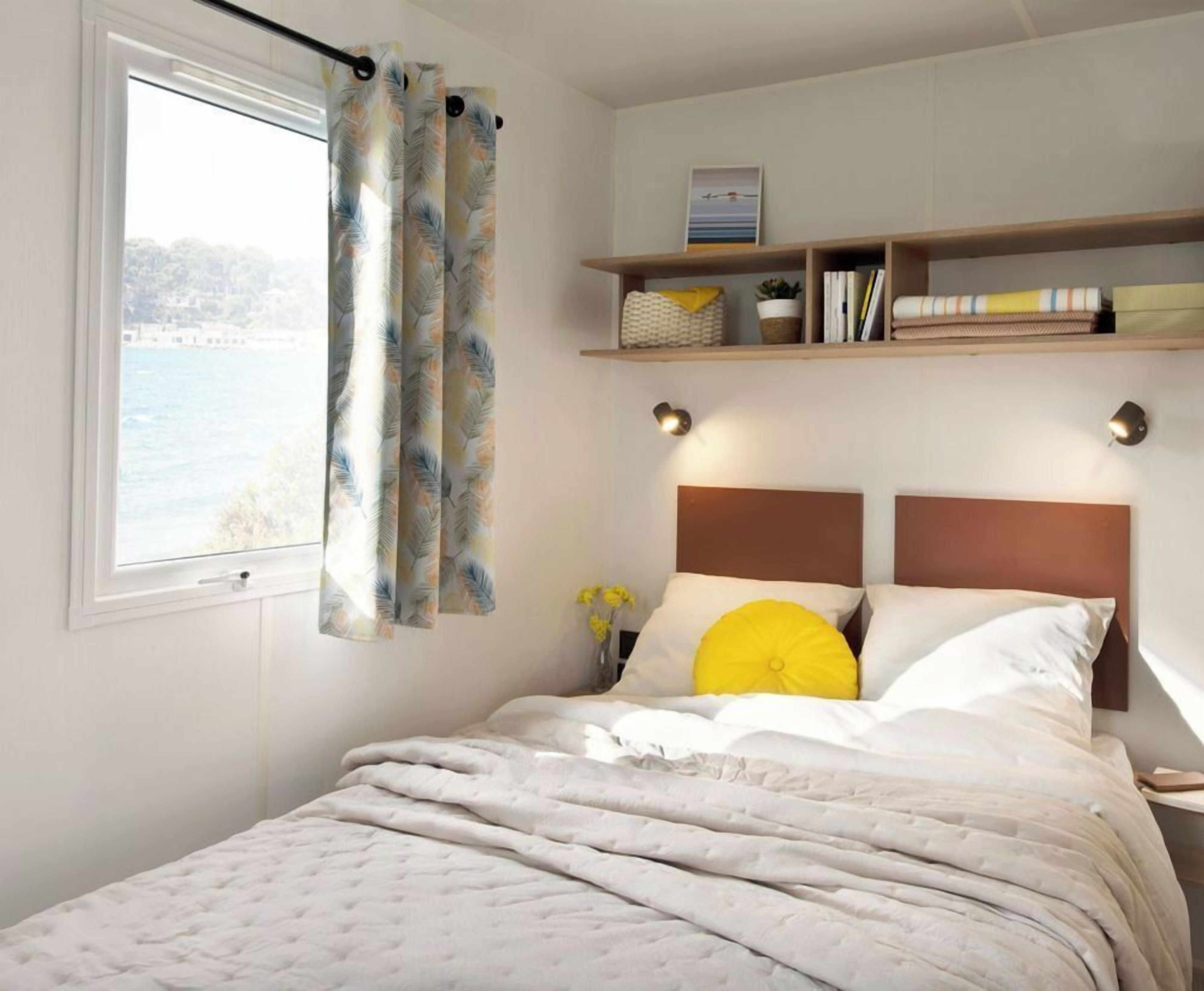 A bedroom with a bed, a window with curtains, a bedside table with a lamp, and a shelf with books and decorative items.