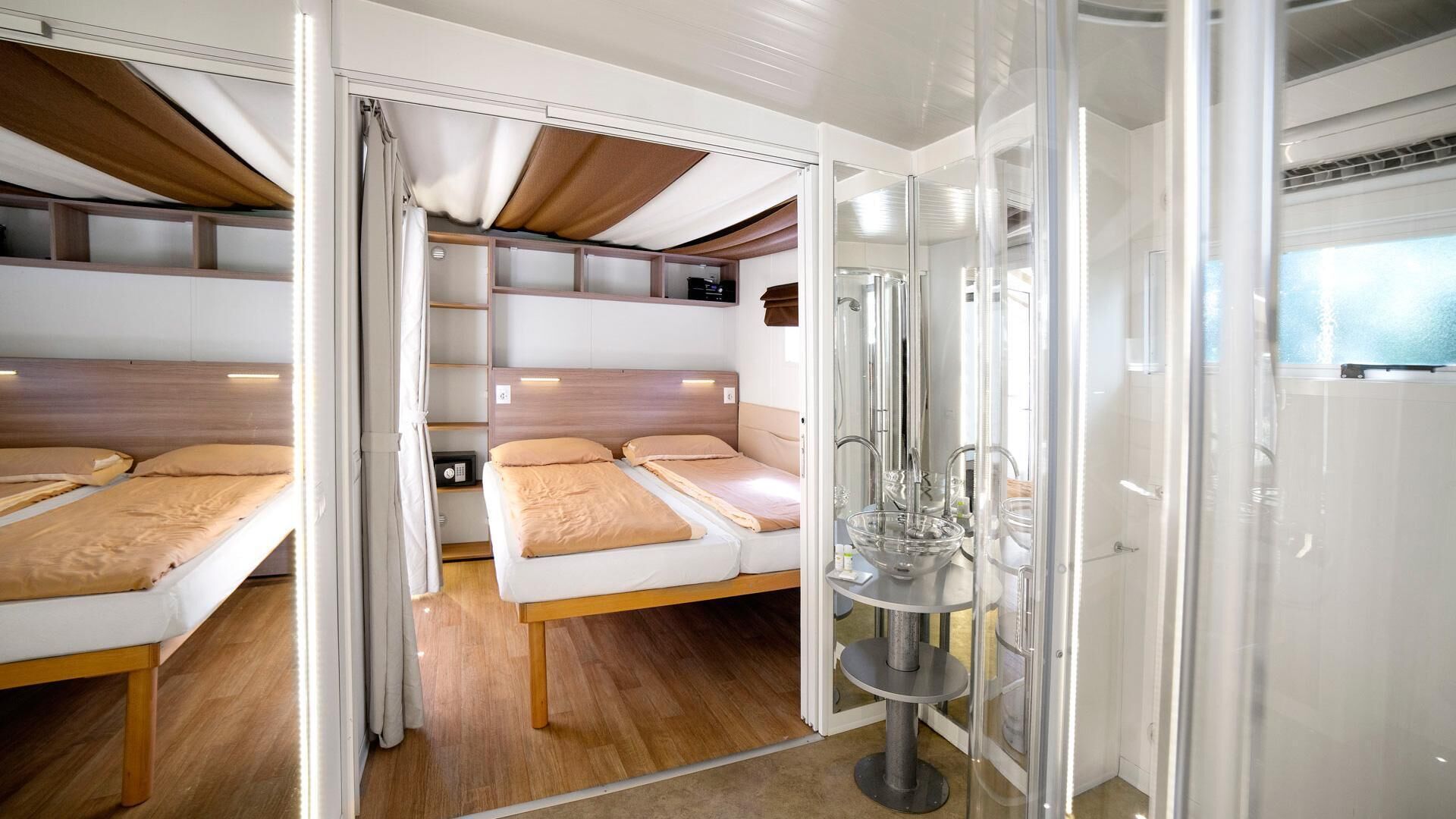 A compact, modern RV interior with two beds, a kitchenette, and a shower area.