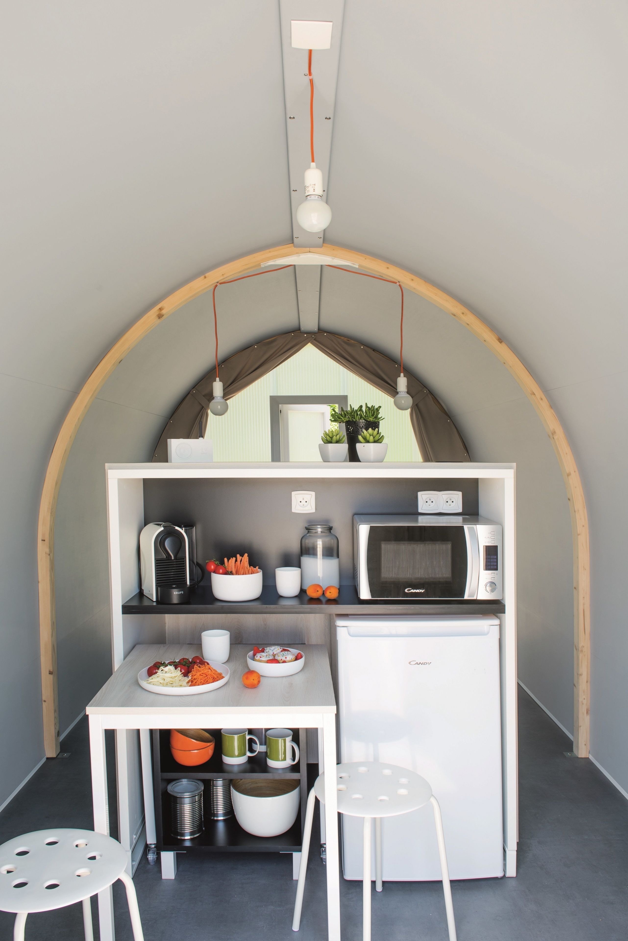 Une petite cuisine équipée d’un four à micro-ondes, d’une bouilloire et d’un petit réfrigérateur. On y trouve une table avec des chaises, une plante en pot et un luminaire suspendu.