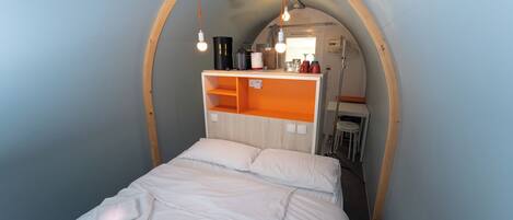 Una habitación pequeña con cama, una mesita y una kitchenette.