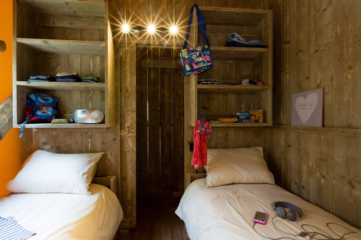Une pièce en bois avec deux lits, une étagère où sont rangés des effets personnels et une porte qui donne sur une autre pièce.