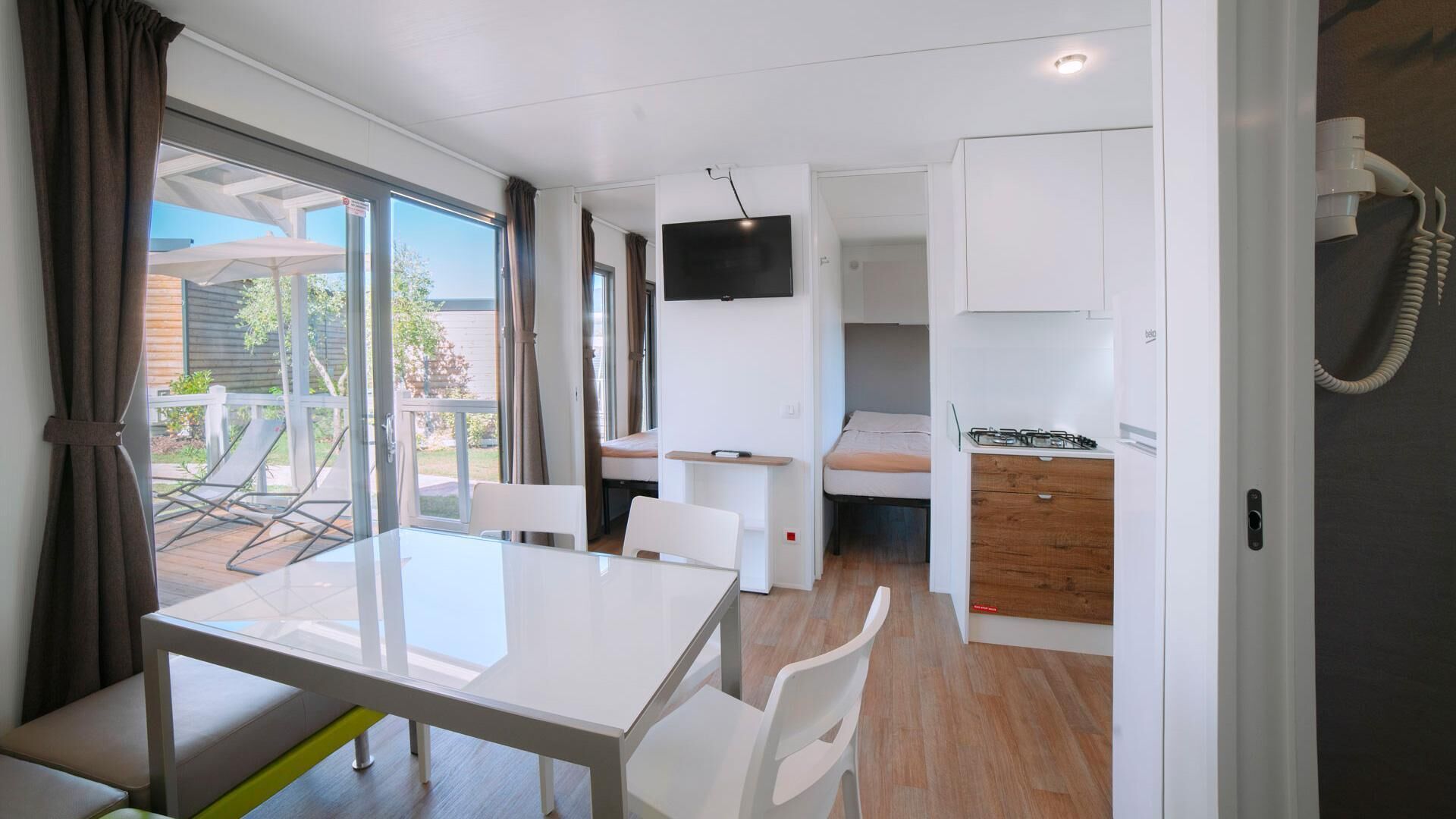 Una habitación compacta con mesa de comedor, sillas, televisor montado en la pared, una kitchenette pequeña y una puerta corrediza que da a un área exterior con patio.