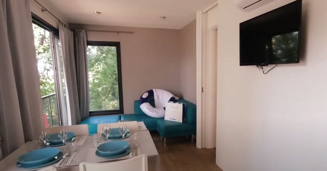 Una sala moderna con un sofá turquesa, un televisor de pantalla plana montado en la pared y una zona de comedor con vajilla azul.