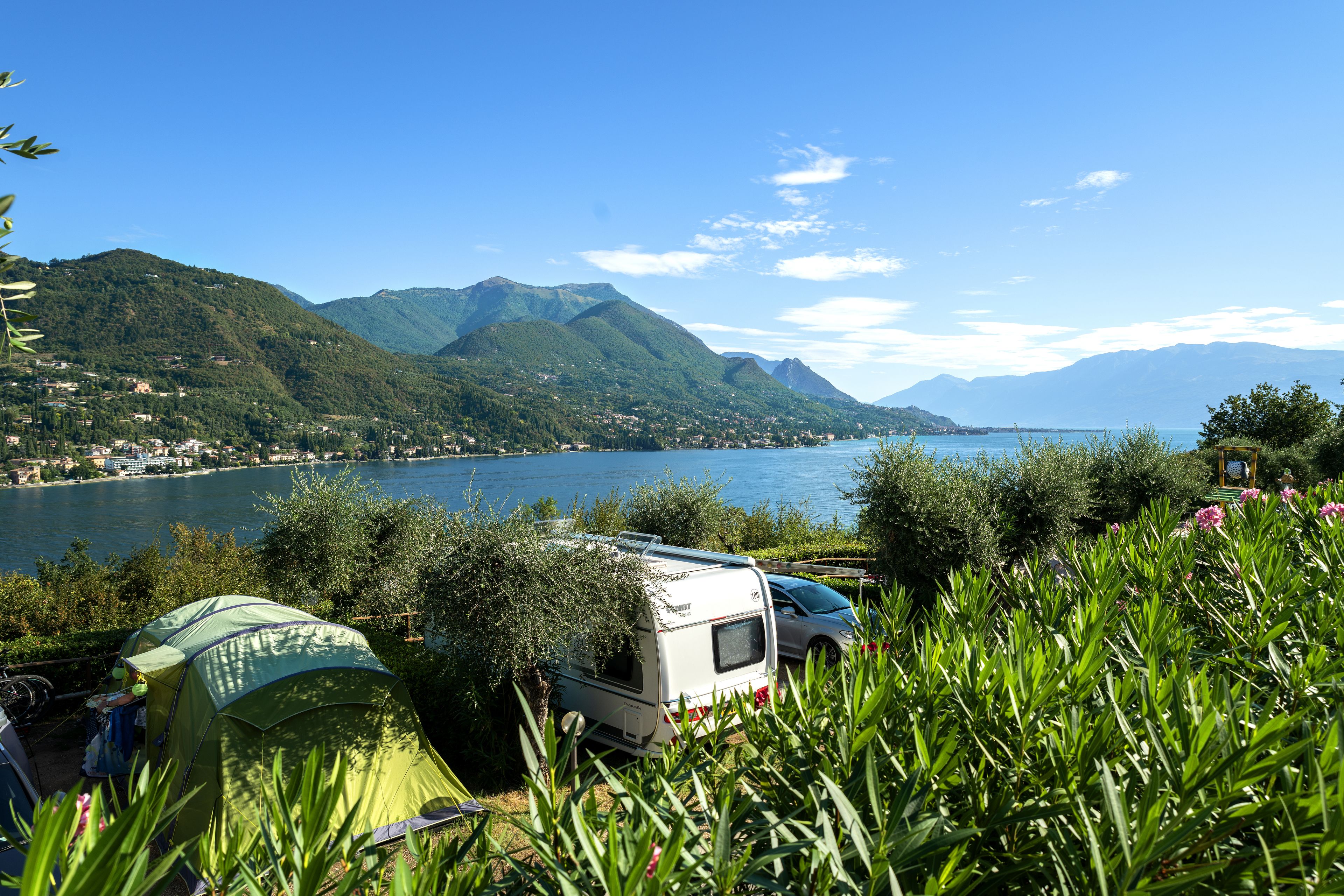 A camping area with a green tent, a white RV, and a lake with mountains in the background.