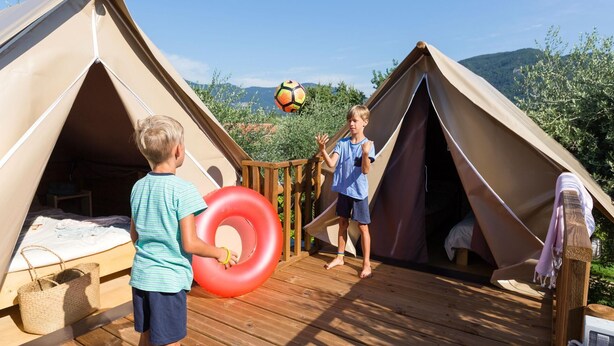 Two children playing with a soccer ball in a tented area with a wooden deck and a view of the hills in the background. - Weekend Glamping Resort (San Felice del Benaco)