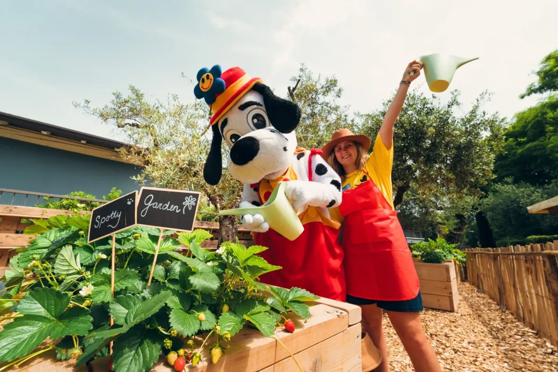 Una persona con un abito rosso tiene un annaffiatoio verde e un cartello con su scritto 'Spotty Garden'.