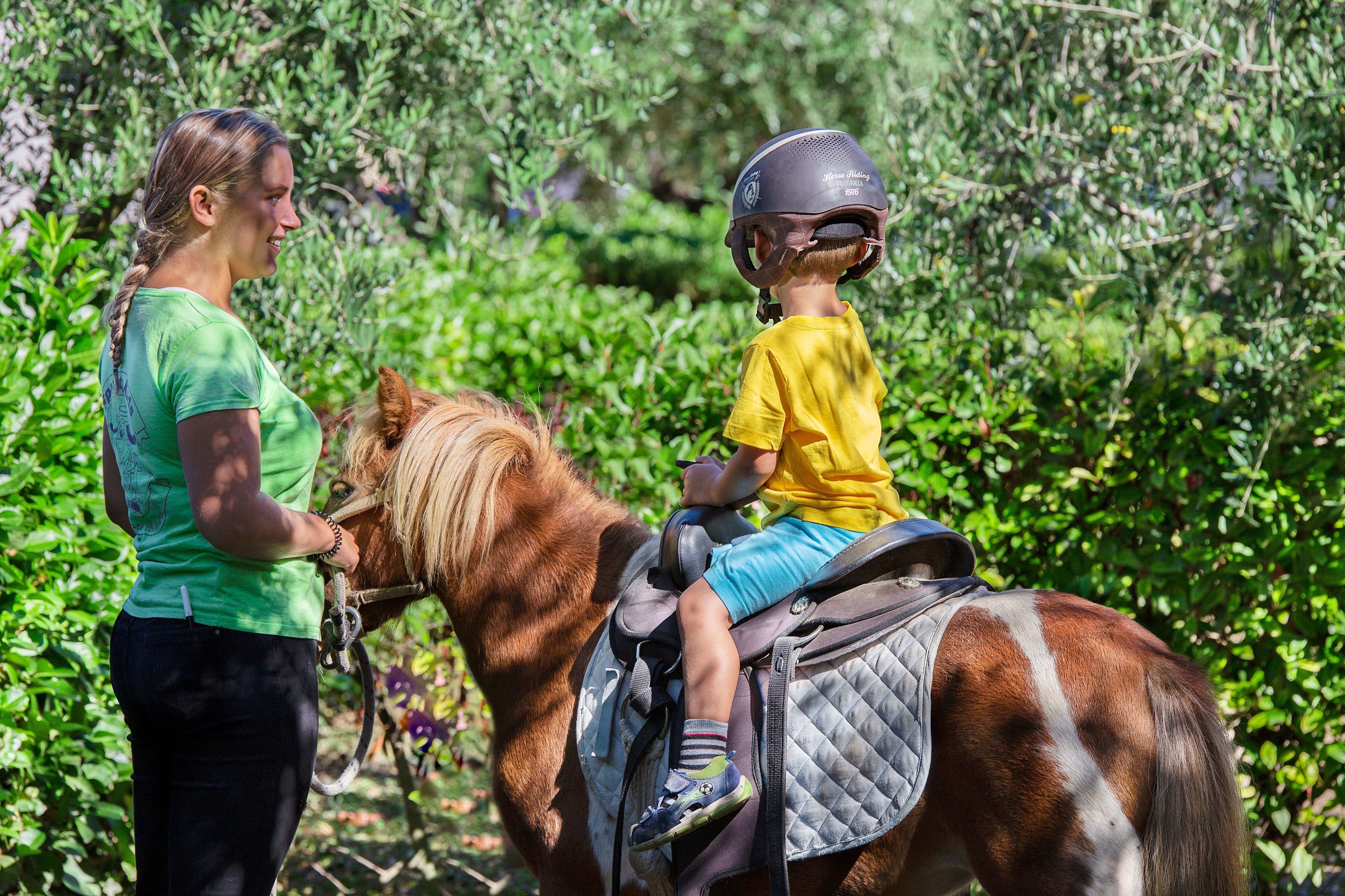 Una persona con camisa verde y pantalones negros está de pie junto a un caballo, sobre el cual viaja un niño con camisa amarilla y pantaloncitos azules.