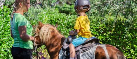Una persona con camisa verde y pantalones negros está de pie junto a un caballo, sobre el cual viaja un niño con camisa amarilla y pantaloncitos azules.