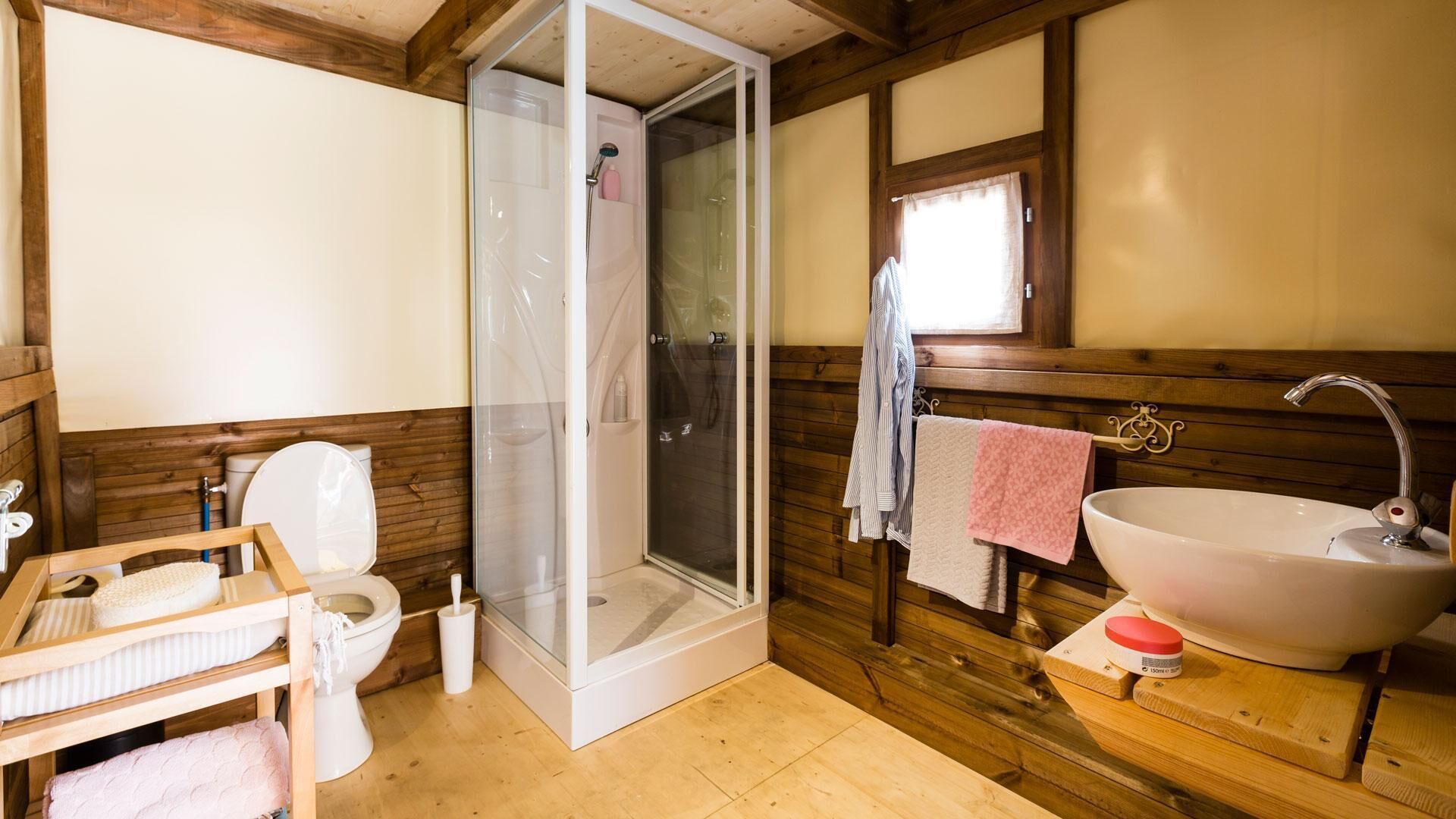 A bathroom with a shower, toilet, and sink, wooden walls, and a window.