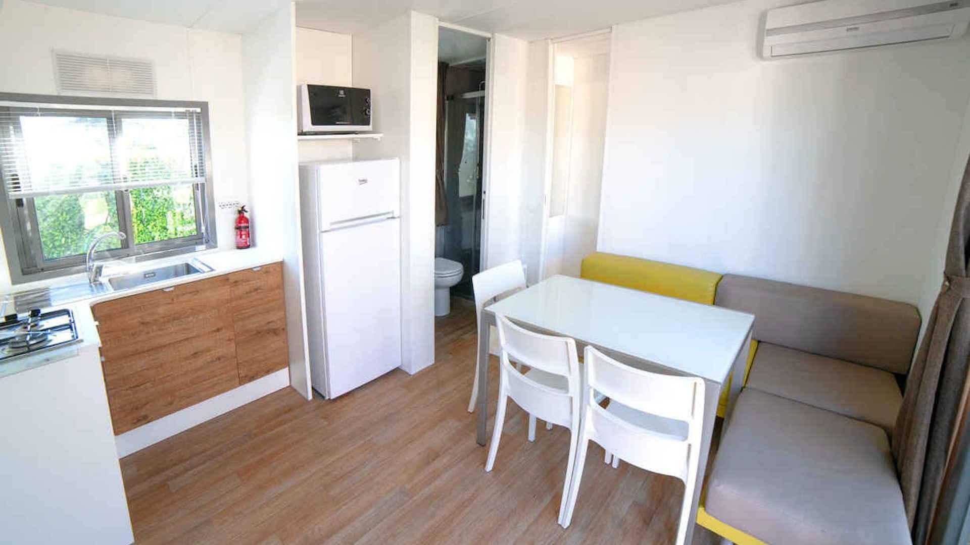 A compact kitchen with a microwave, sink, and a refrigerator. There is a dining table with chairs and a sofa with cushions.