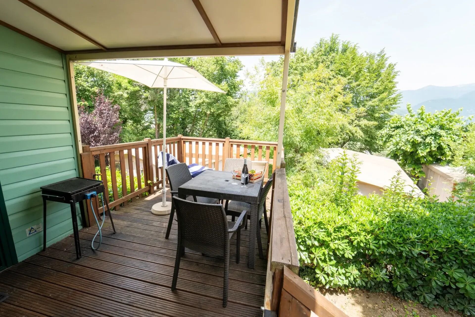 En overdækket terrasse med et bord og stole, grill og udsigt til grøn natur og bjerge.