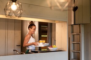 Una mujer con camiseta blanca y delantal verde lleva una bandeja con jugo de naranja y un bol de fruta.