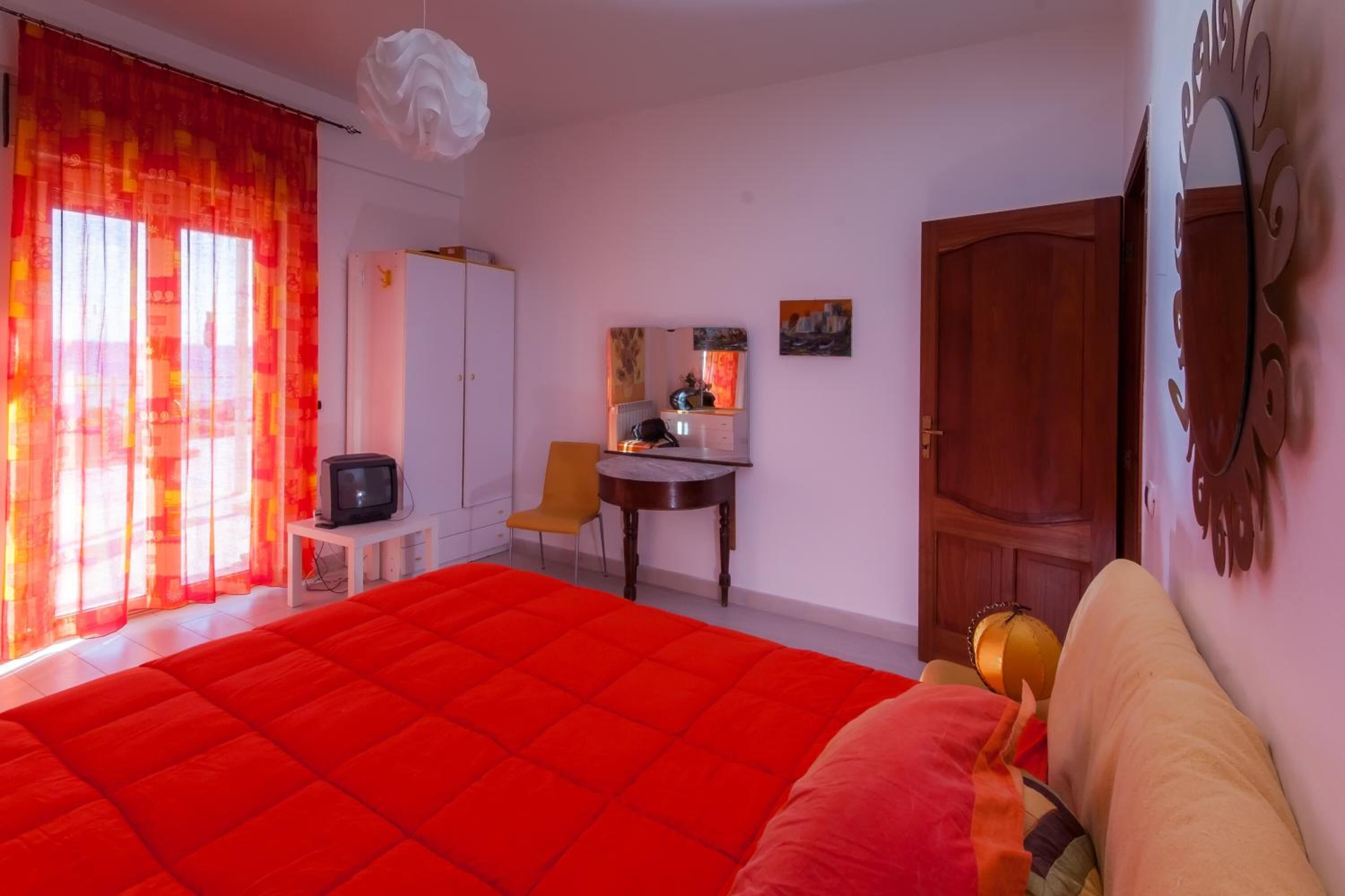 Un dormitorio con una cama roja grande, una puerta de madera, una mesita con un televisor y una silla.