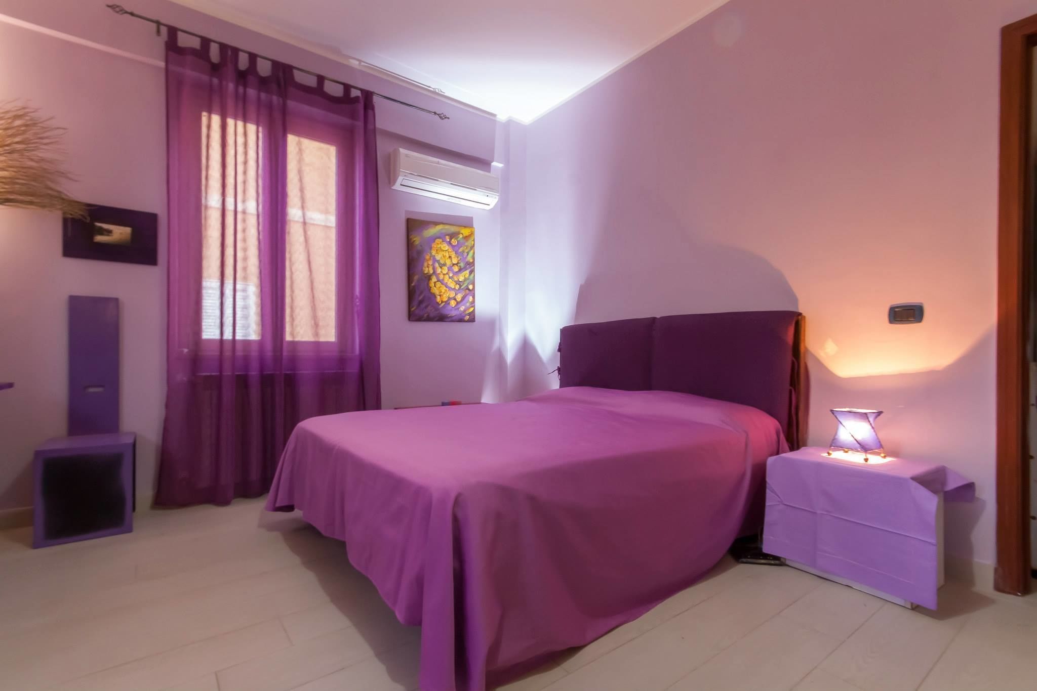 Ein Schlafzimmer mit einem Bett, einem Nachttisch, einer Lampe, einem Fenster mit violetten Vorhängen und einer Wand-Klimaanlage.