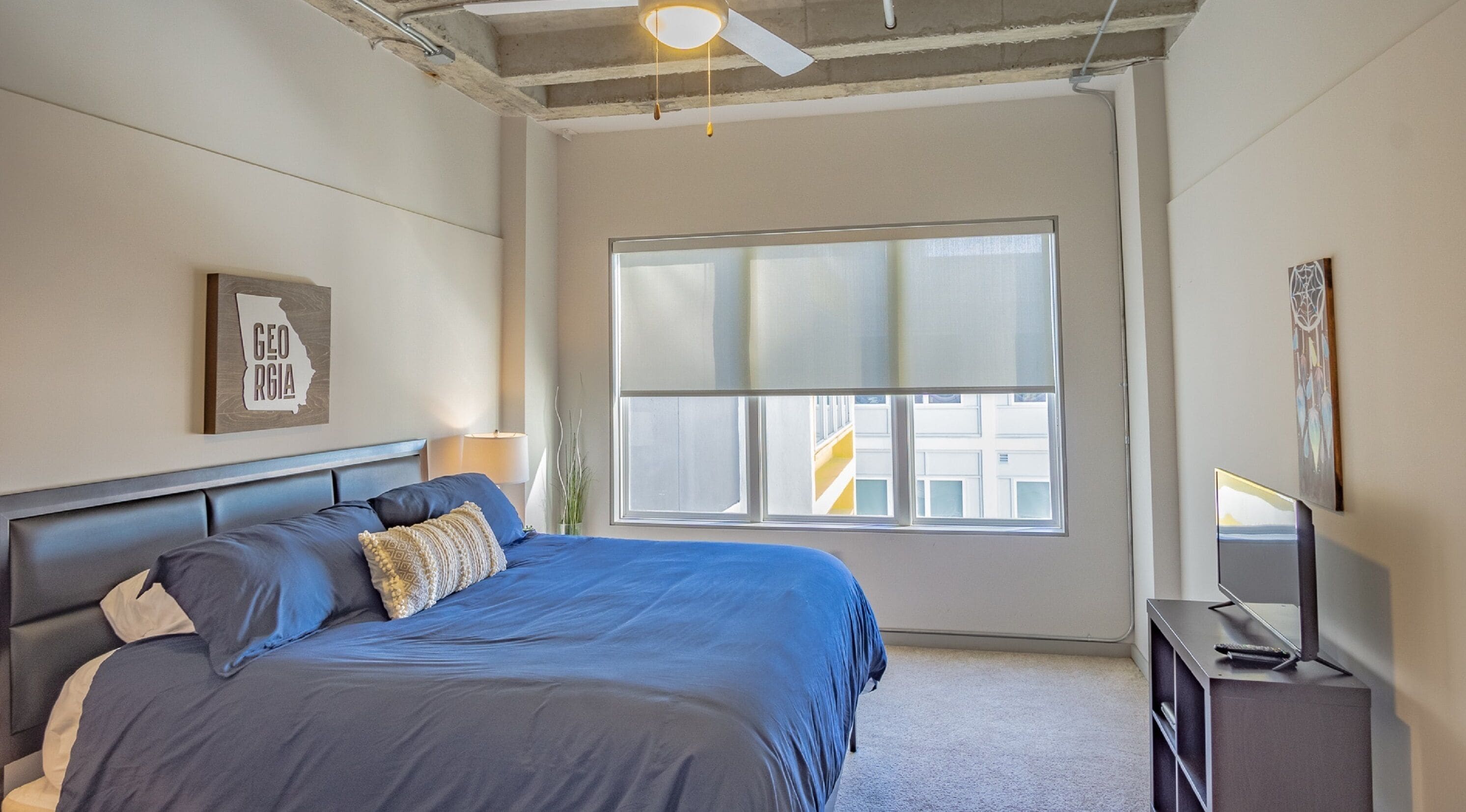 A bedroom with a bed, a window, a ceiling fan, and a TV stand.