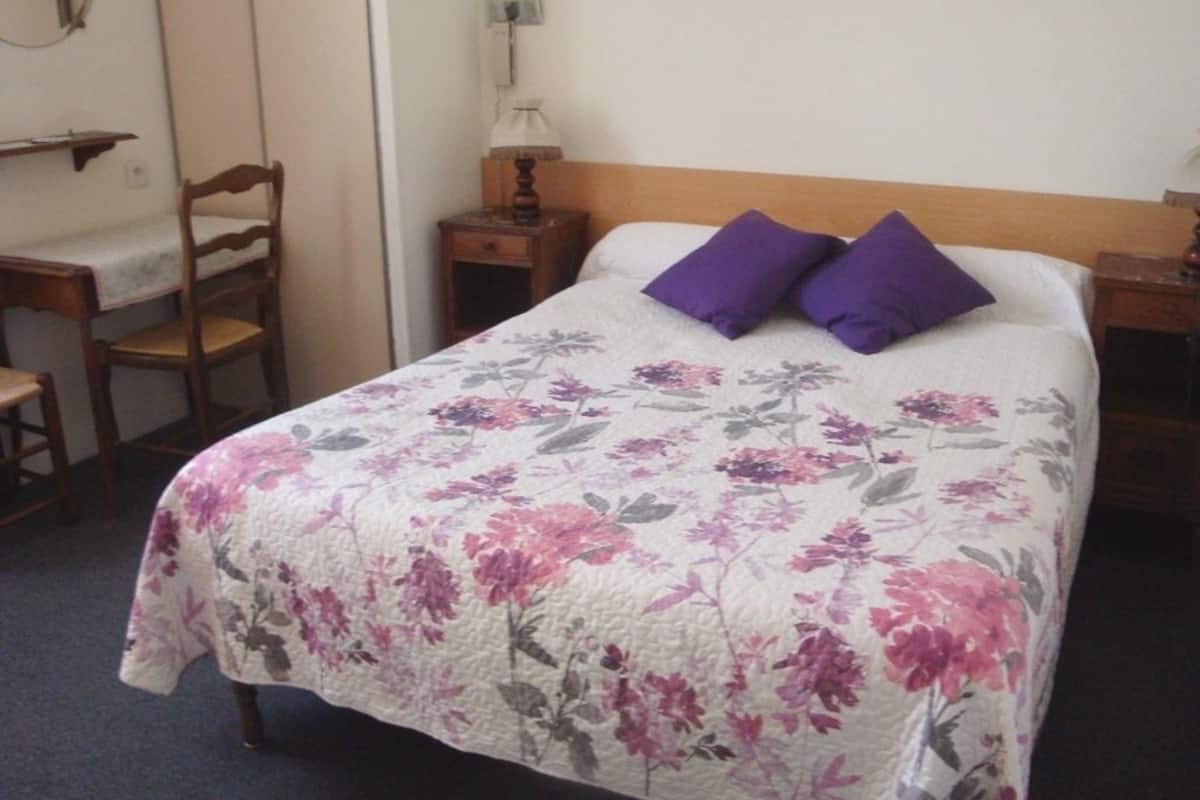 A bed with a floral bedspread, two purple pillows, a wooden headboard, a nightstand with a lamp, and a chair with a table.