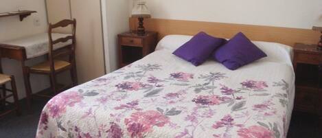 A bed with a floral bedspread, two purple pillows, a wooden headboard, a nightstand with a lamp, and a chair with a table.