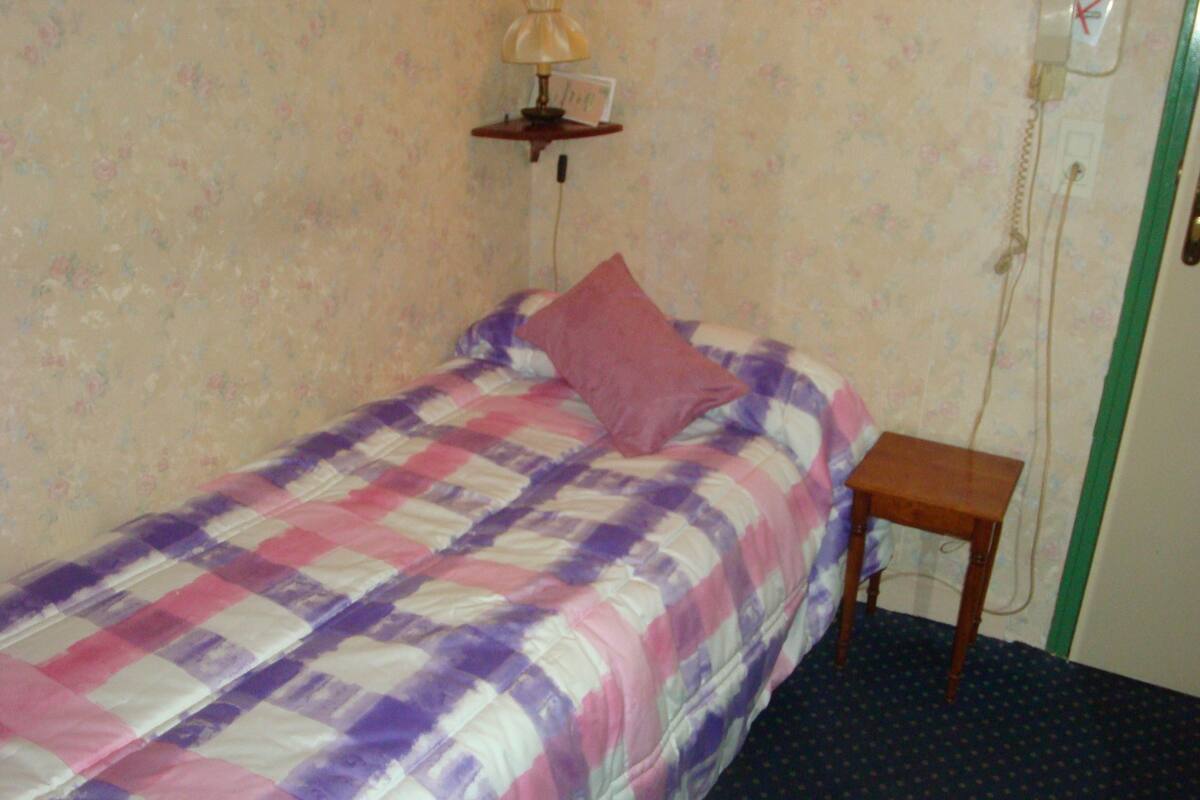 A single bed with a checkered bedspread, a bedside table, a lamp, and a telephone on the wall.