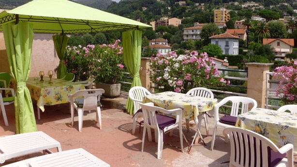 An outdoor terrace with a table and chairs, a green canopy, and a view of a hillside with houses. - Hôtel La Lubiane (Vence)