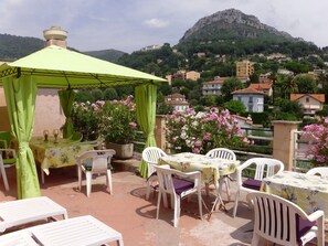 An outdoor terrace with a table and chairs, a green canopy, and a view of a hillside with houses. - Hôtel La Lubiane (Vence)