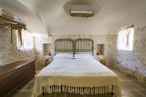 A bedroom with a stone wall, a wooden bench, a bed with a green metal frame, and a white air conditioner mounted on the wall.