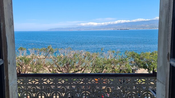 Vista desde un balcón con barandilla, sobre un cuerpo de agua y montañas a lo lejos.