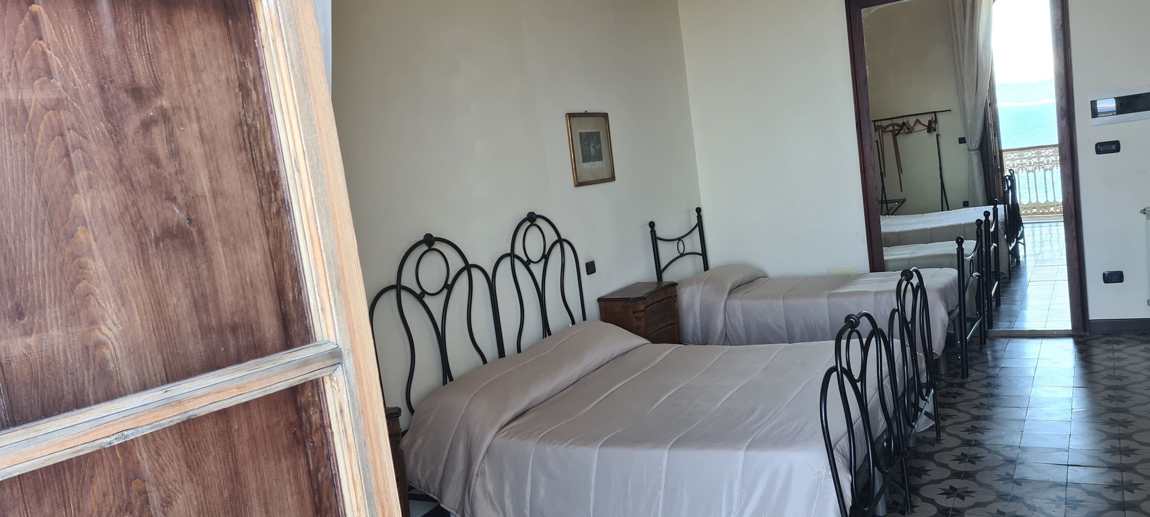 A room with two beds, a wooden door, a small wooden nightstand, a metal headboard, a framed picture, and a view of the sea through a window.