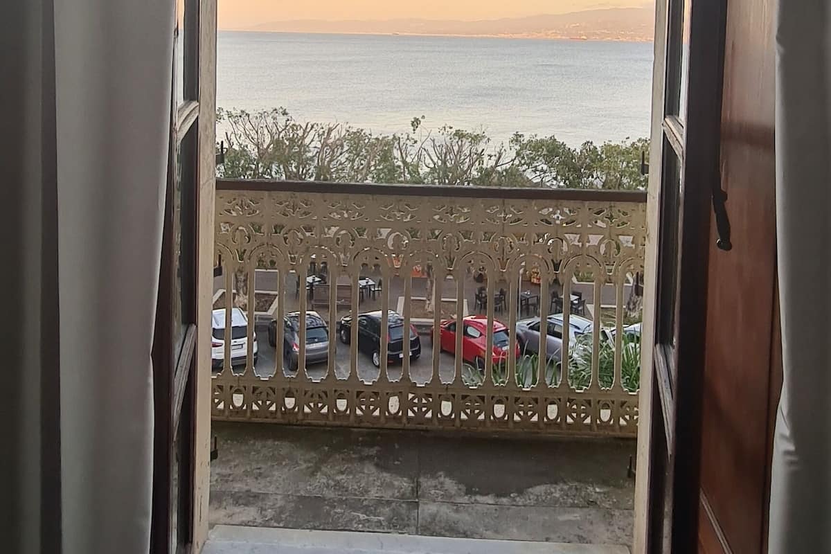 An open doorway leading to a balcony with a view of the sea and parked cars.