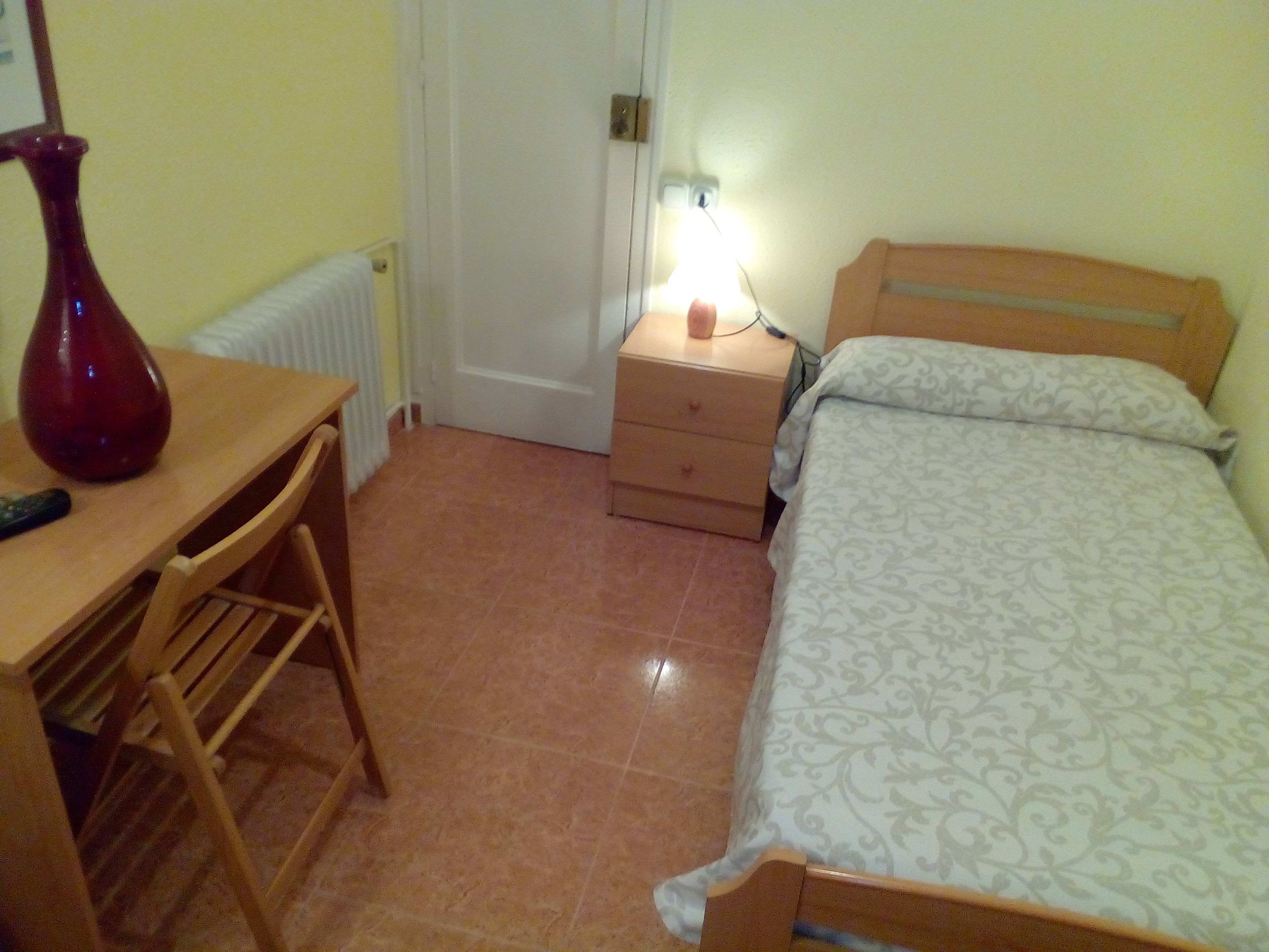 Quarto com cama, mesa de cabeceira, luminária, cadeira e escrivaninha de madeira com um vaso.