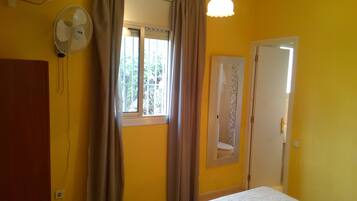 Una habitación con cama, ventilador, ventana con cortinas, puerta y lámpara.