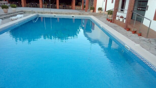 A rectangular swimming pool with a tiled edge, surrounded by a patio area with steps leading up to a building entrance. - Hostal Lleida (Calafell)