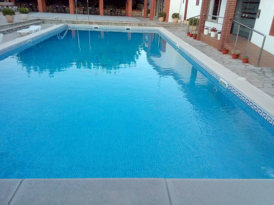 Piscina rectangular con borde de azulejos, rodeada por una terraza con escaleras que conducen a la entrada de un edificio.