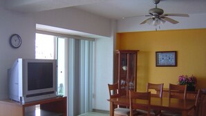 A dining area with a wooden table and chairs, a television on a stand, a ceiling fan, and a clock on the wall.
