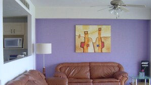 A living room with a brown leather sofa, a glass coffee table with a red bowl, a ceiling fan, a wall-mounted painting, and a microwave in a cabinet.