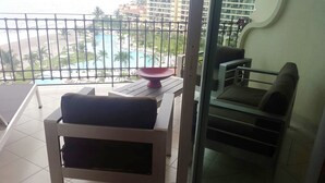 A balcony with a view of a pool and palm trees, a dining table with chairs, and a sofa.