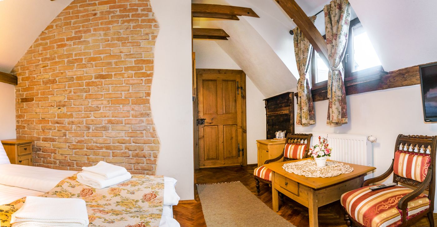 A cozy room with a brick wall, wooden beams, a bed with floral bedding, a wooden table with chairs, and a window with curtains.