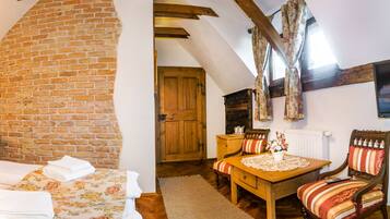 A cozy room with a brick wall, wooden beams, a bed with floral bedding, a wooden table with chairs, and a window with curtains.