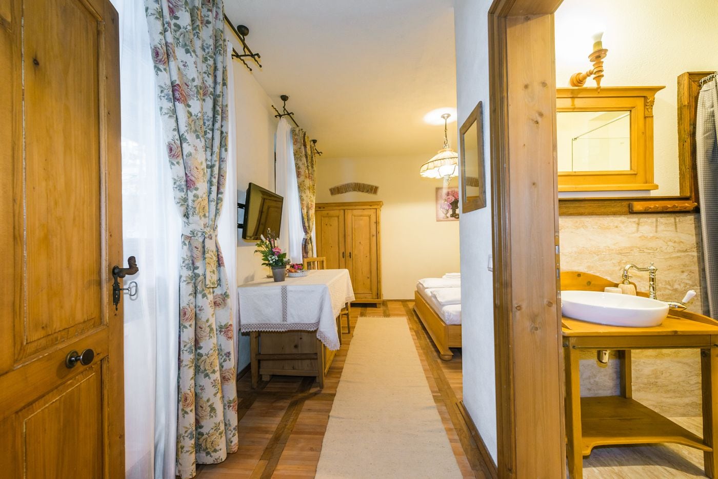 Un pasillo estrecho que conduce a un dormitorio con una puerta de madera, una mesa con mantel floral y una cama con ropa blanca.