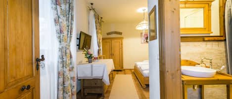 Un pasillo estrecho que conduce a un dormitorio con una puerta de madera, una mesa con mantel floral y una cama con ropa blanca.