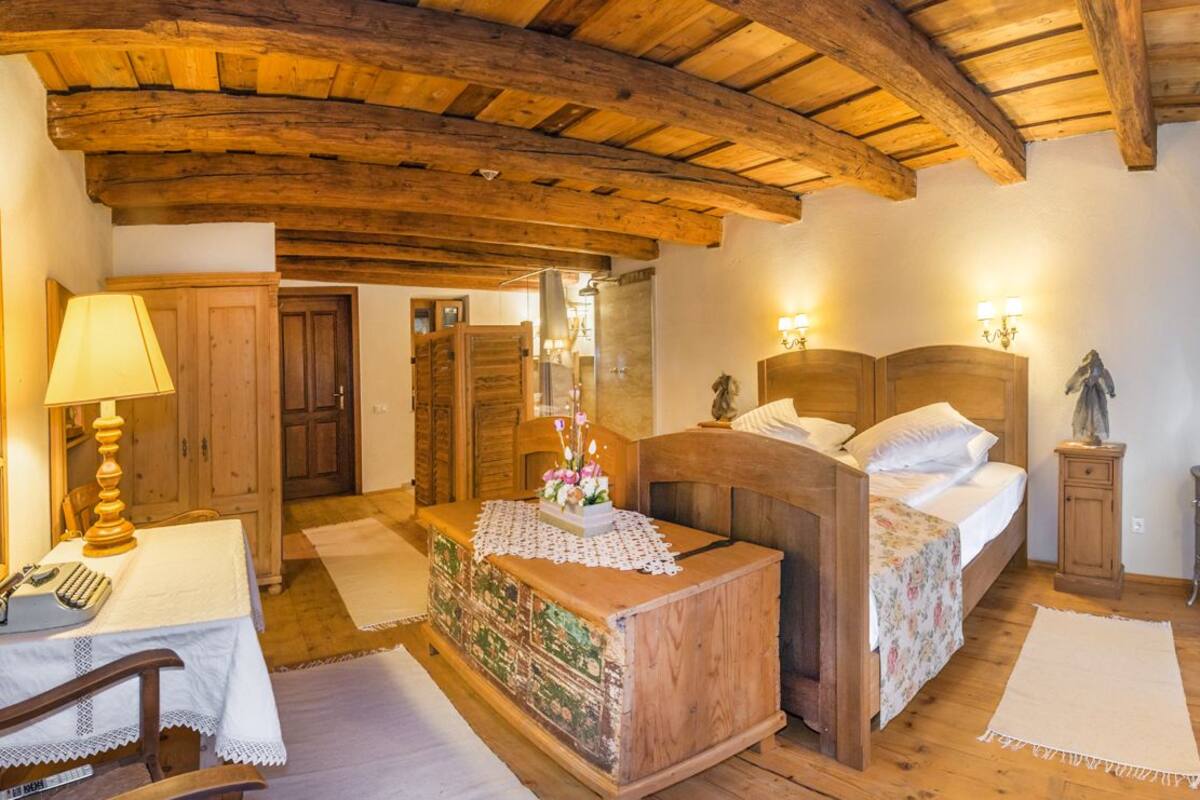Una habitación rústica con vigas de madera, una cama con sábanas blancas, un escritorio con lámpara y un aparador con un arreglo floral.