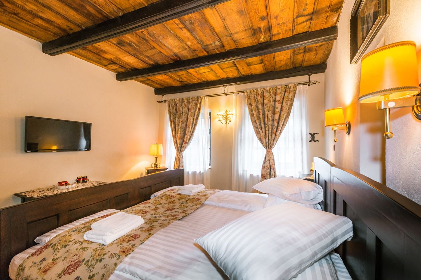 Une chambre d’hôtel avec un plafond en bois, un lit recouvert d’une literie à motifs floraux, deux lampes de chevet et un téléviseur à écran plat.