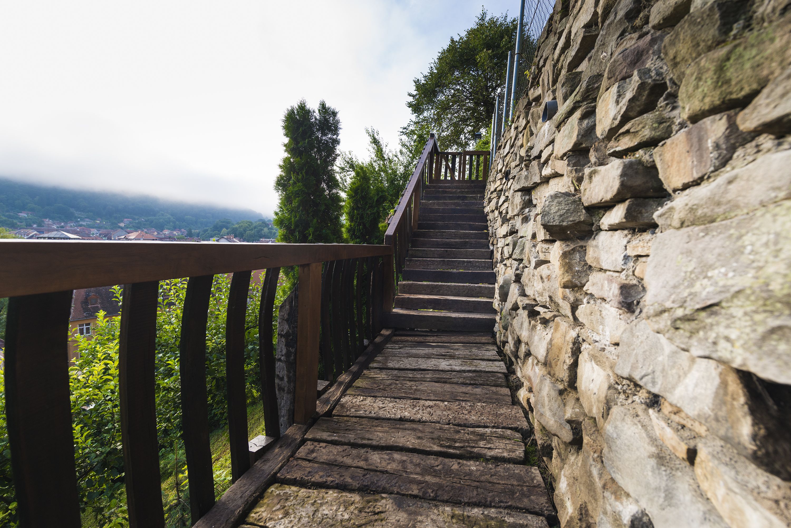A stone staircase with a wooden railing leading up a hill, surrounded by greenery and overlooking a town.