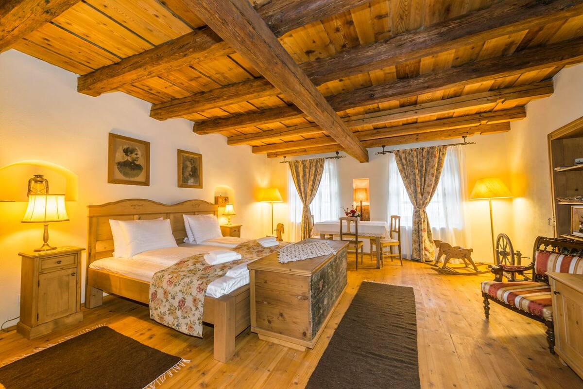 Una habitación acogedora con techo de madera, una cama con colcha estampada, un cofre de madera, un escritorio con una silla y una lámpara antigua.